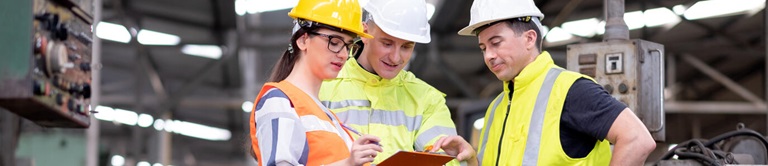 Three Caucasian factory workers or technician discuss together in front of the other employee in workplace area in safety gear